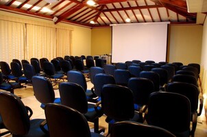 Un salón de conferencias con hileras de sillas negras, una pantalla de proyección y un techo de madera.