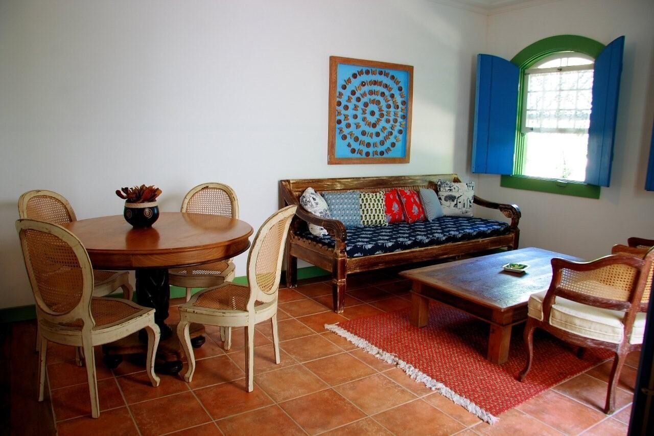 A room with a wooden dining table, wicker chairs, a sofa with patterned cushions, a red rug, and a window with blue shutters.