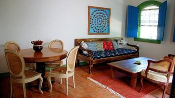 A room with a wooden dining table, wicker chairs, a sofa with patterned cushions, a red rug, and a window with blue shutters.