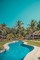 Una piscina de borde curvo, rodeada de palmeras y una estructura con techo de paja.