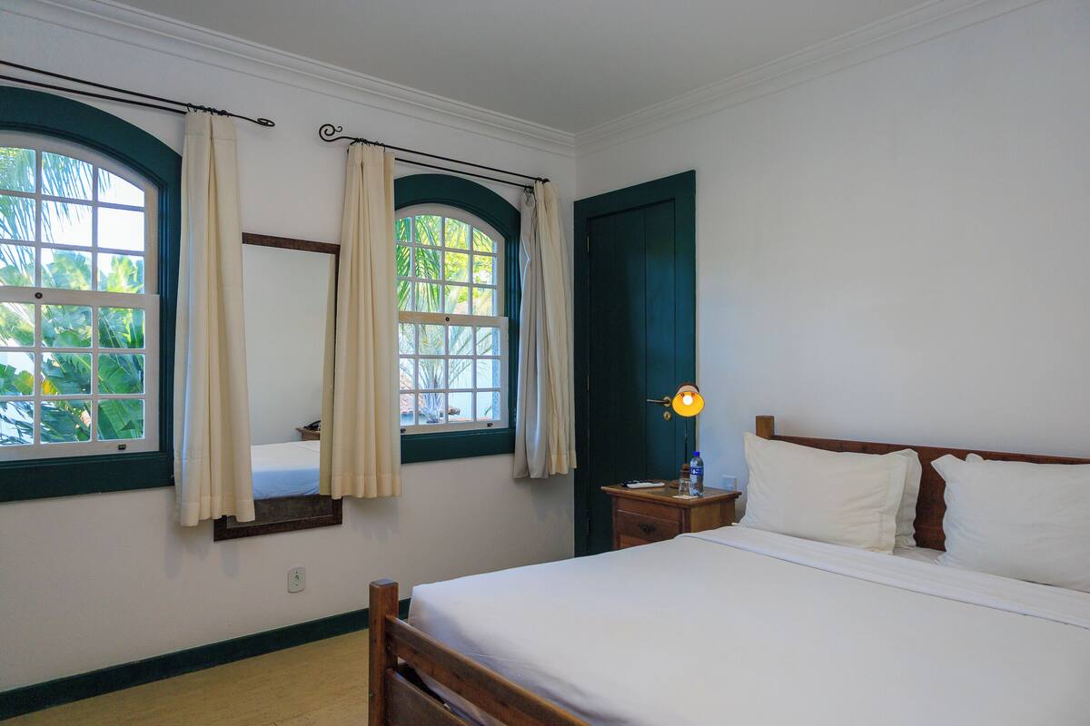 Un dormitorio con una cama, dos ventanas con cortinas, una mesita de noche con lámpara y un cabecero de madera.