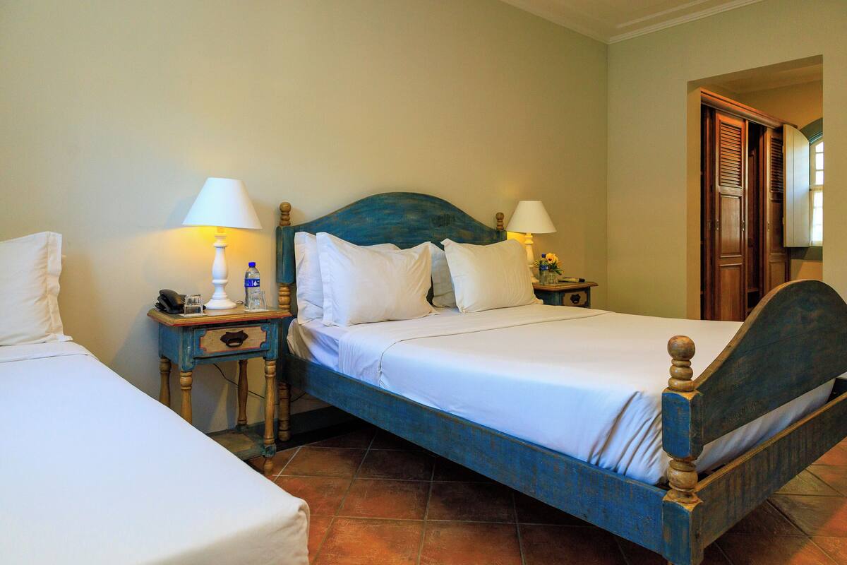 Una cama doble con cabecera de madera, dos mesitas de noche con lámparas y un mueble auxiliar con un teléfono y una botella.