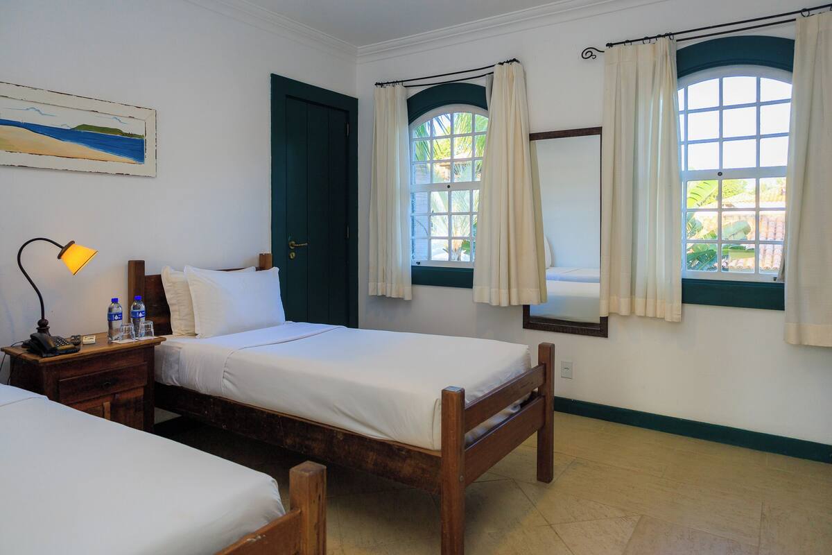 Habitación de hotel con dos camas, una mesita de noche con lámpara, un cuadro y una ventana con cortinas.