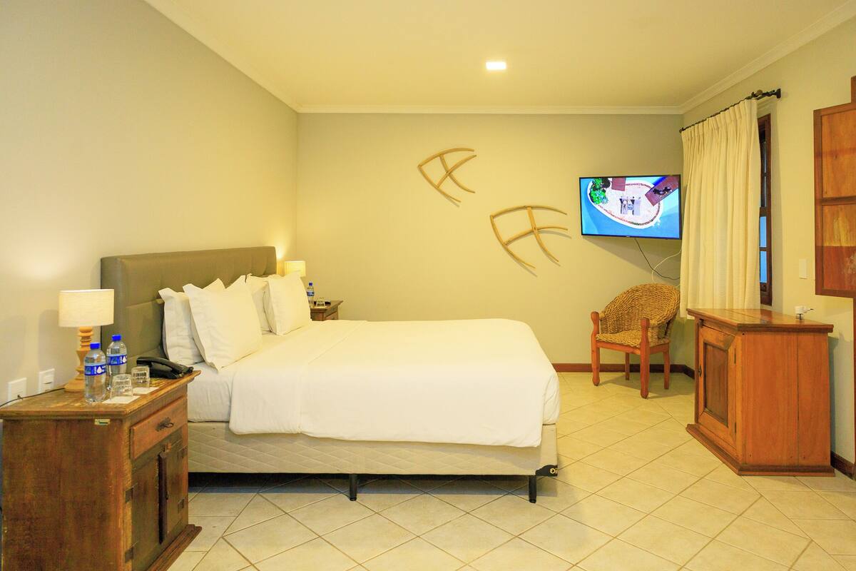 Una habitación de hotel con cama, una mesita de noche de madera con lámpara, un televisor montado en la pared y una silla.