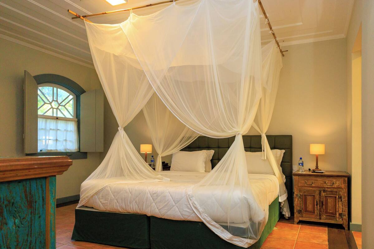 Una cama con dosel y cuatro columnas, una mesita de noche con lámpara, una ventana con vistas y un armario de madera.