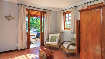 A room with a wooden cabinet, wicker chairs with cushions, a wooden chest of drawers, and a view through a glass door to an outdoor area with a table and chairs.