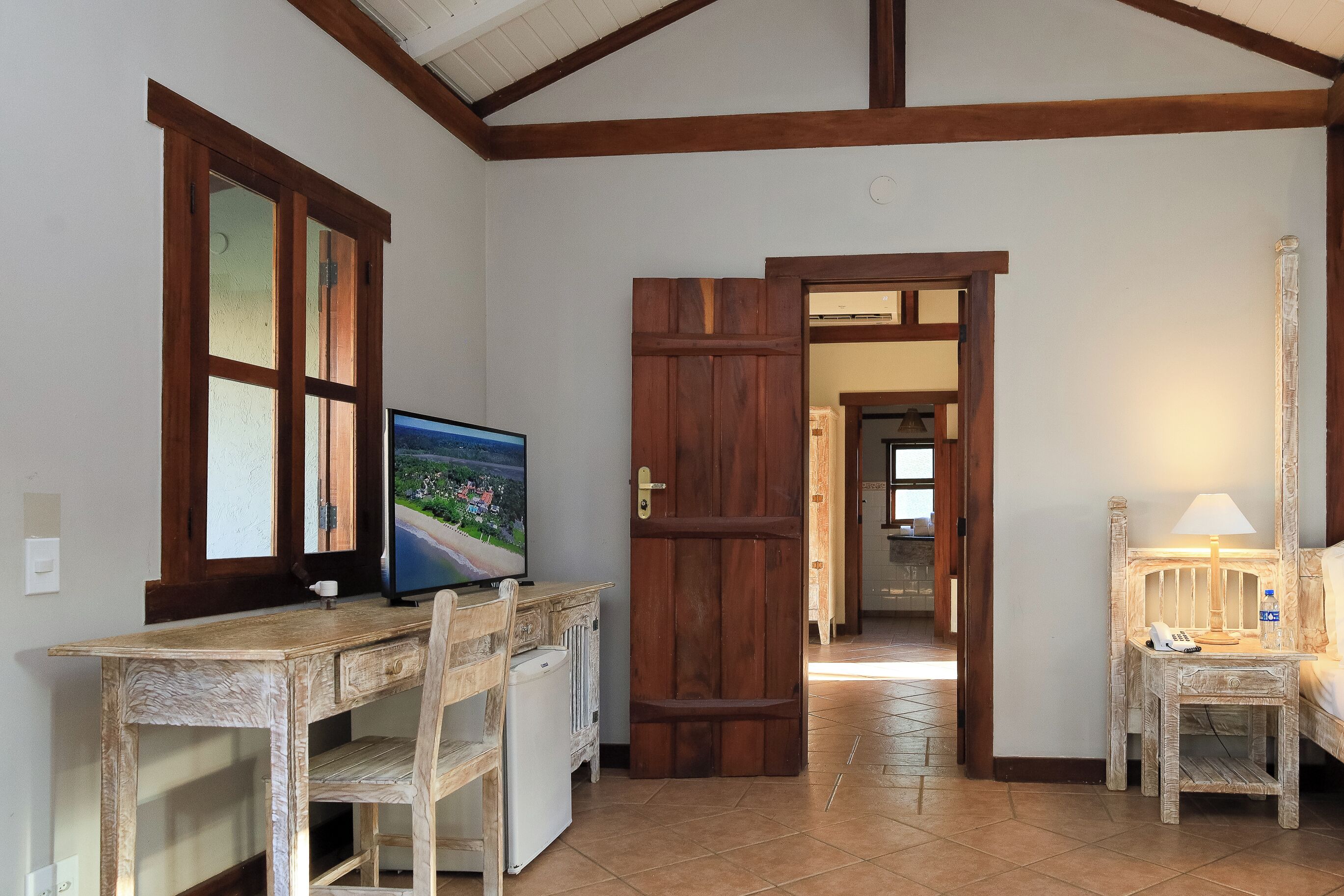 Una habitación con un escritorio de madera, una televisión, una cama con mesita de noche y una puerta que da a otra estancia.