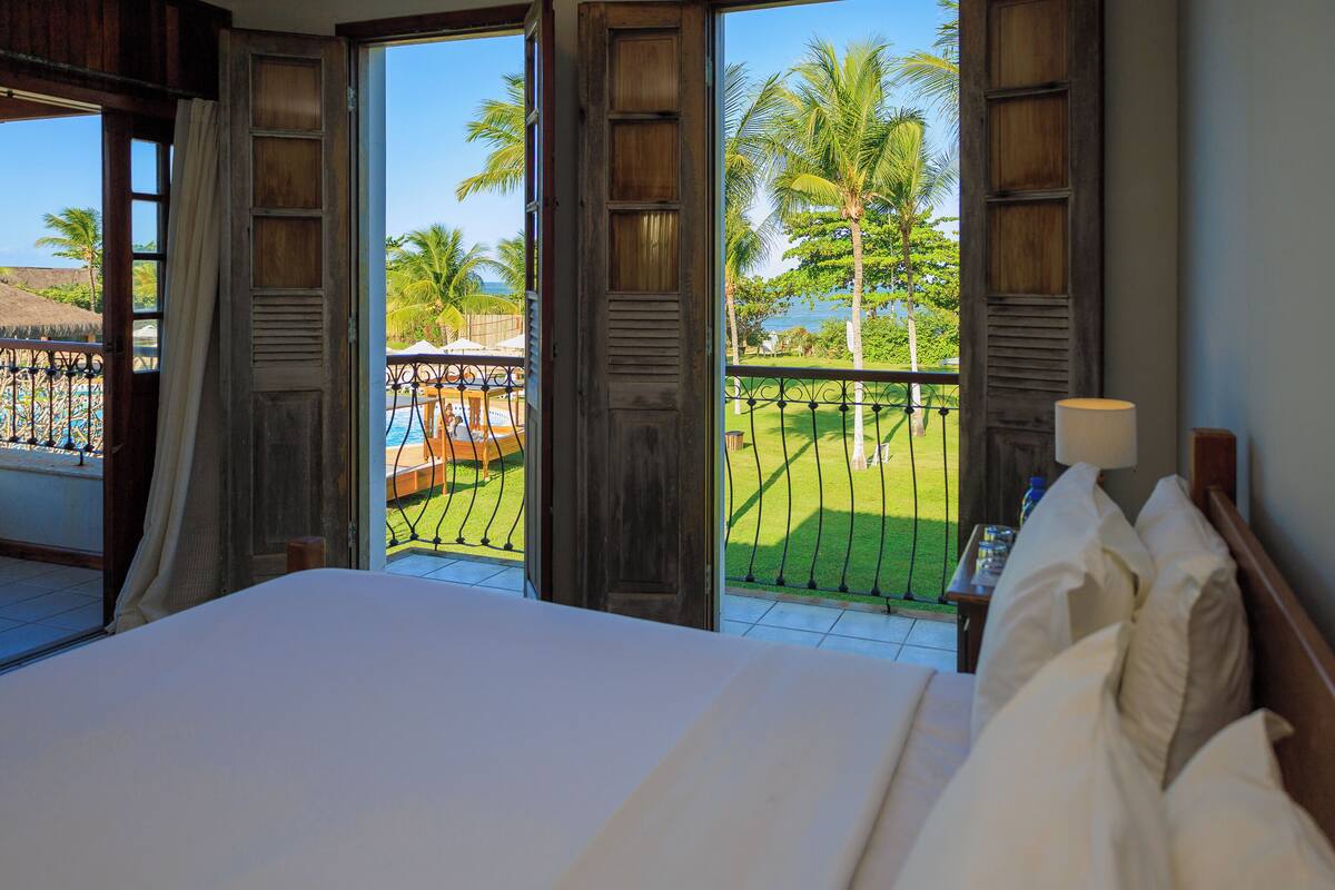 Habitación de hotel con una cama grande, vista a una piscina y palmeras, y un balcón con barandilla.