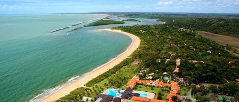 An aerial view of a coastal resort with a beach, swimming pools, and a mix of residential and commercial buildings.