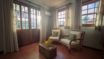 A room with two wicker chairs, a small wooden table with a vase, and large windows with curtains.
