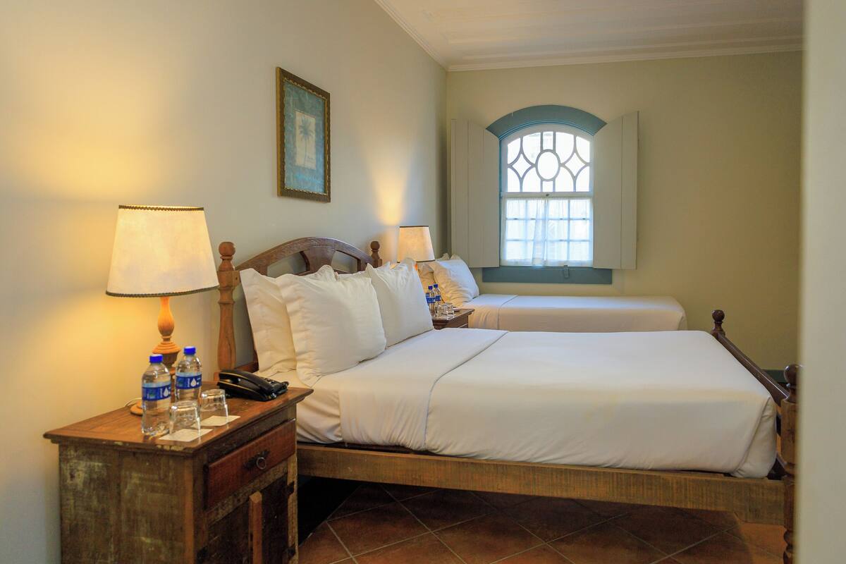 Habitación de hotel con dos camas, una mesita de noche con lámpara, un cuadro enmarco y una ventana con arco decorativo.