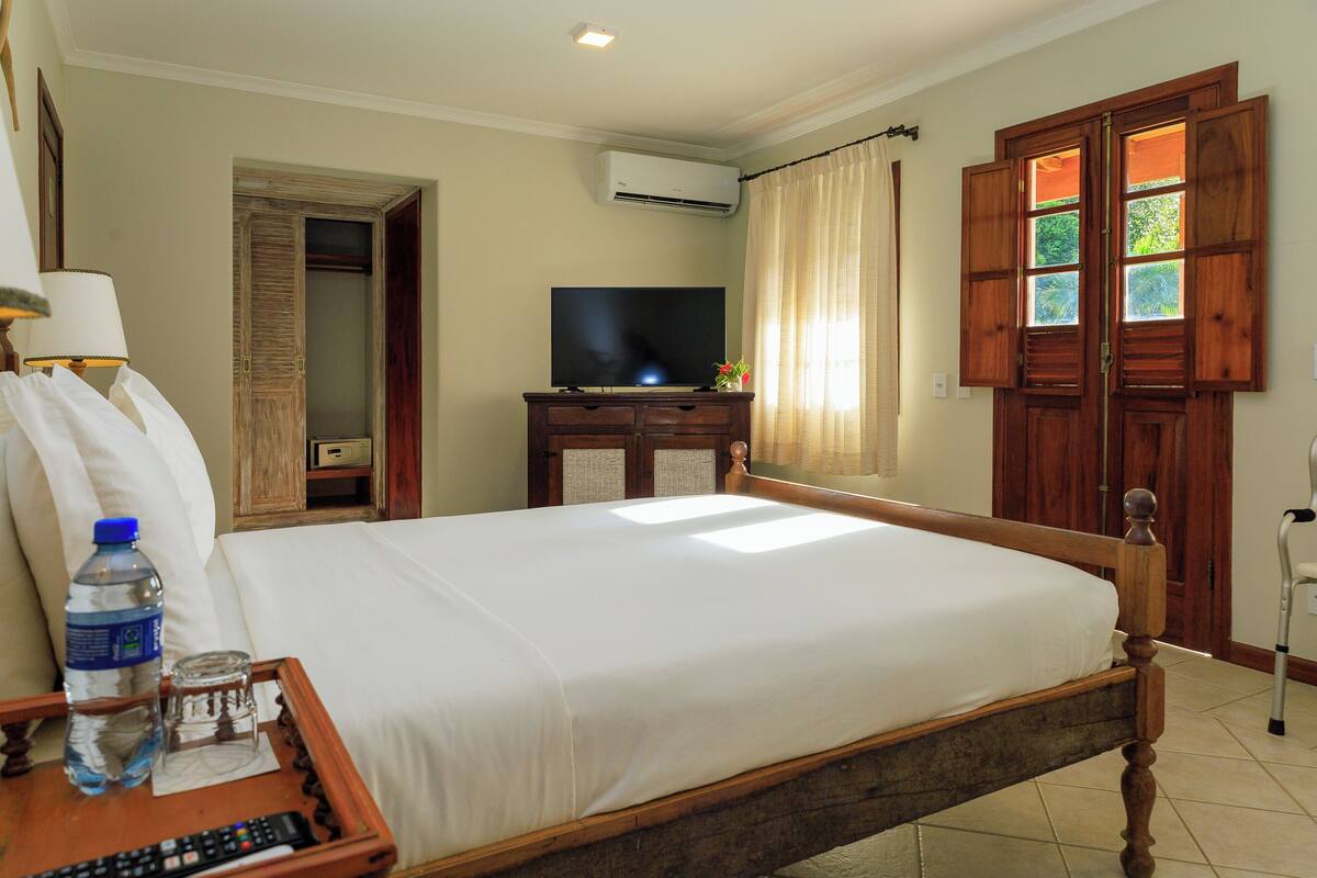 Habitación de hotel con cama, televisión, botella de agua, vaso y control remoto sobre una bandeja de madera.