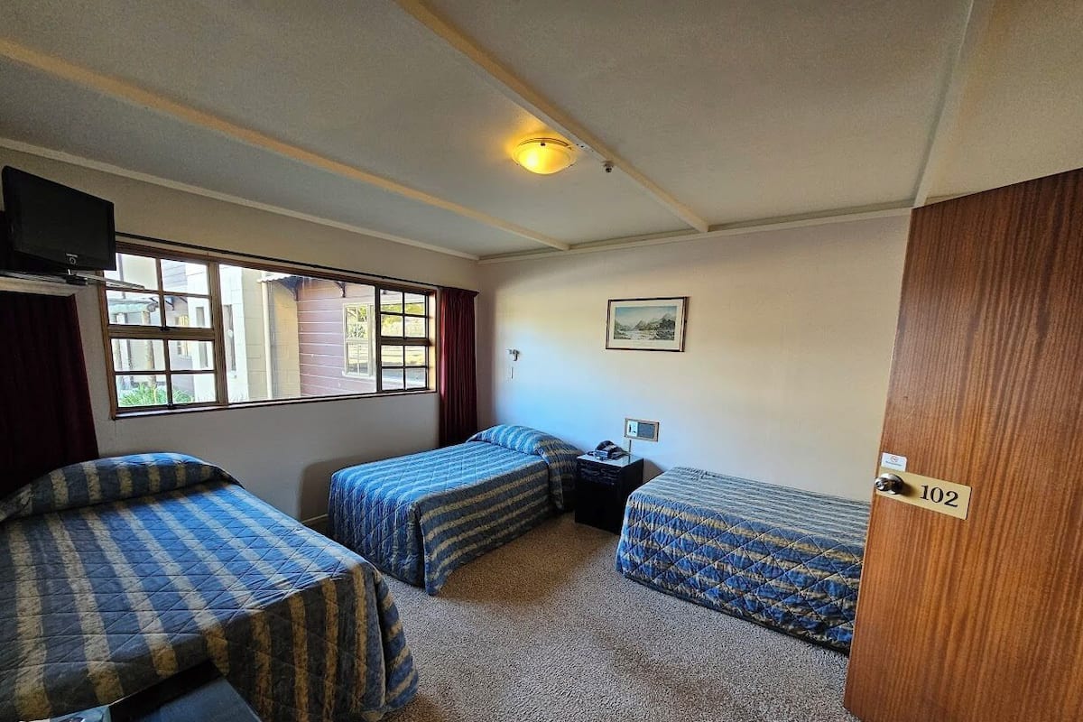 A room with three beds, a desk, a chair, a nightstand, a lamp, a window, and a door with a number plate.