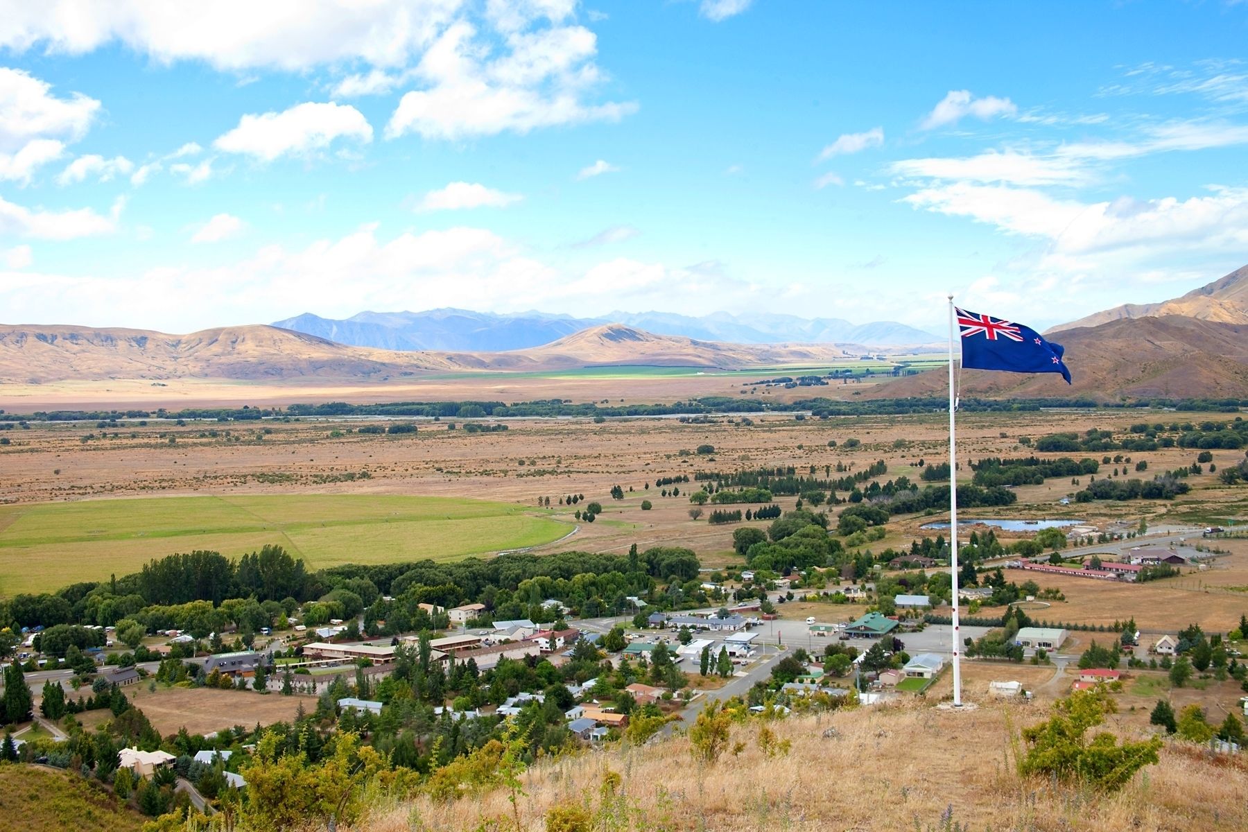 A flag on a flagpole in a rural town with houses and mountains in the background.