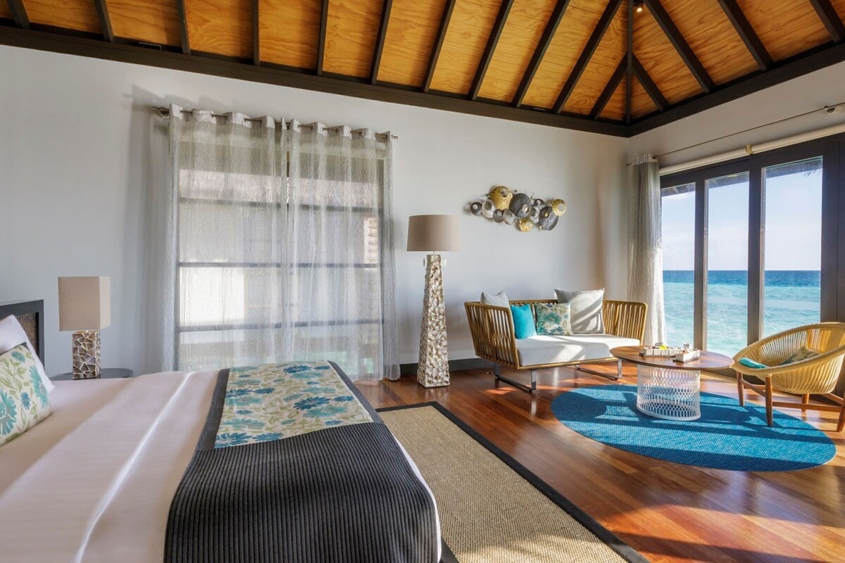 A hotel room with a bed, a sofa, a chair, a lamp, and a view of the sea.