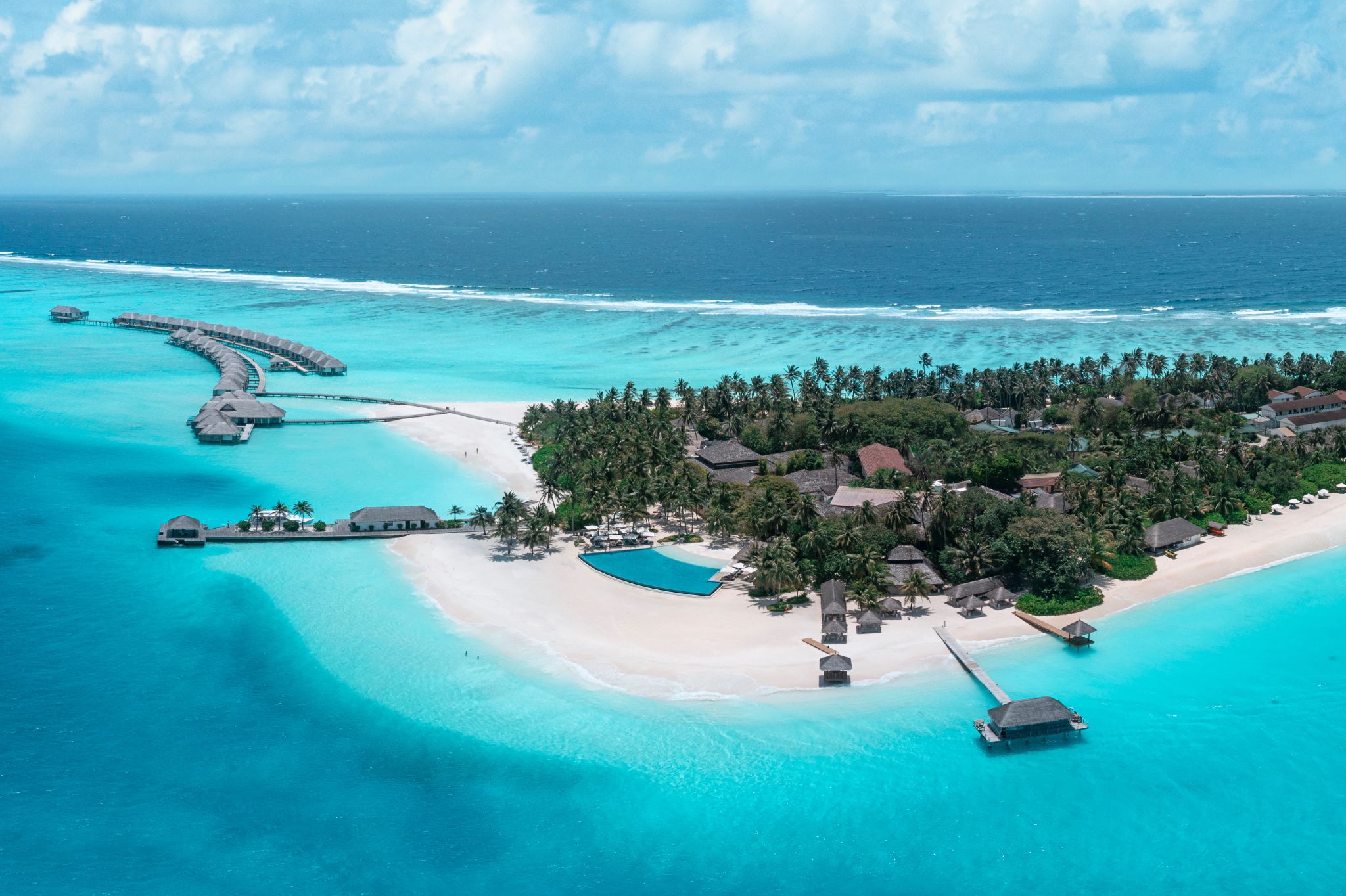 An aerial view of a tropical resort with overwater bungalows, a beach, and a clear blue sea.