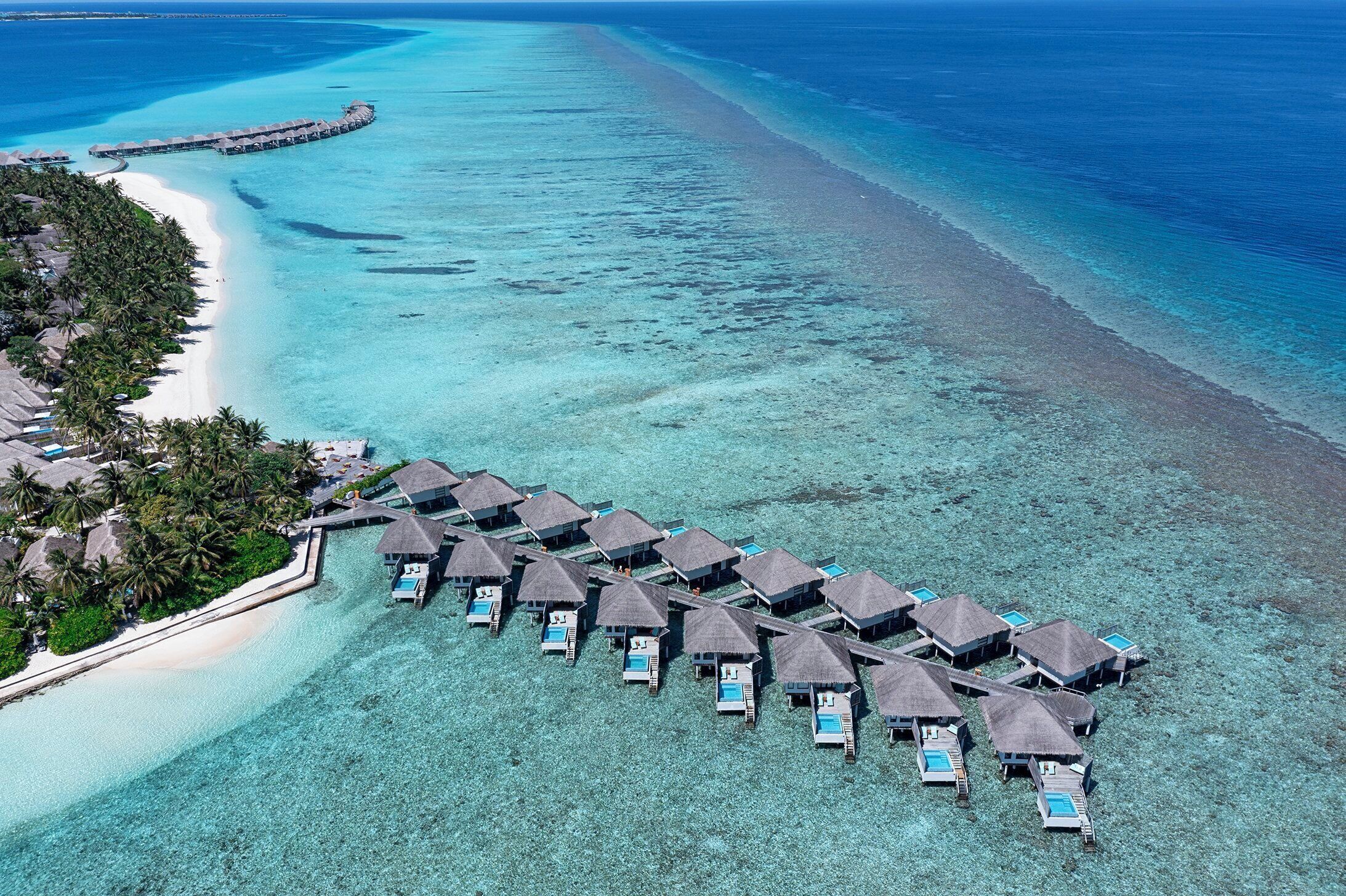 An aerial view of overwater bungalows with clear blue water and a sandy beach.