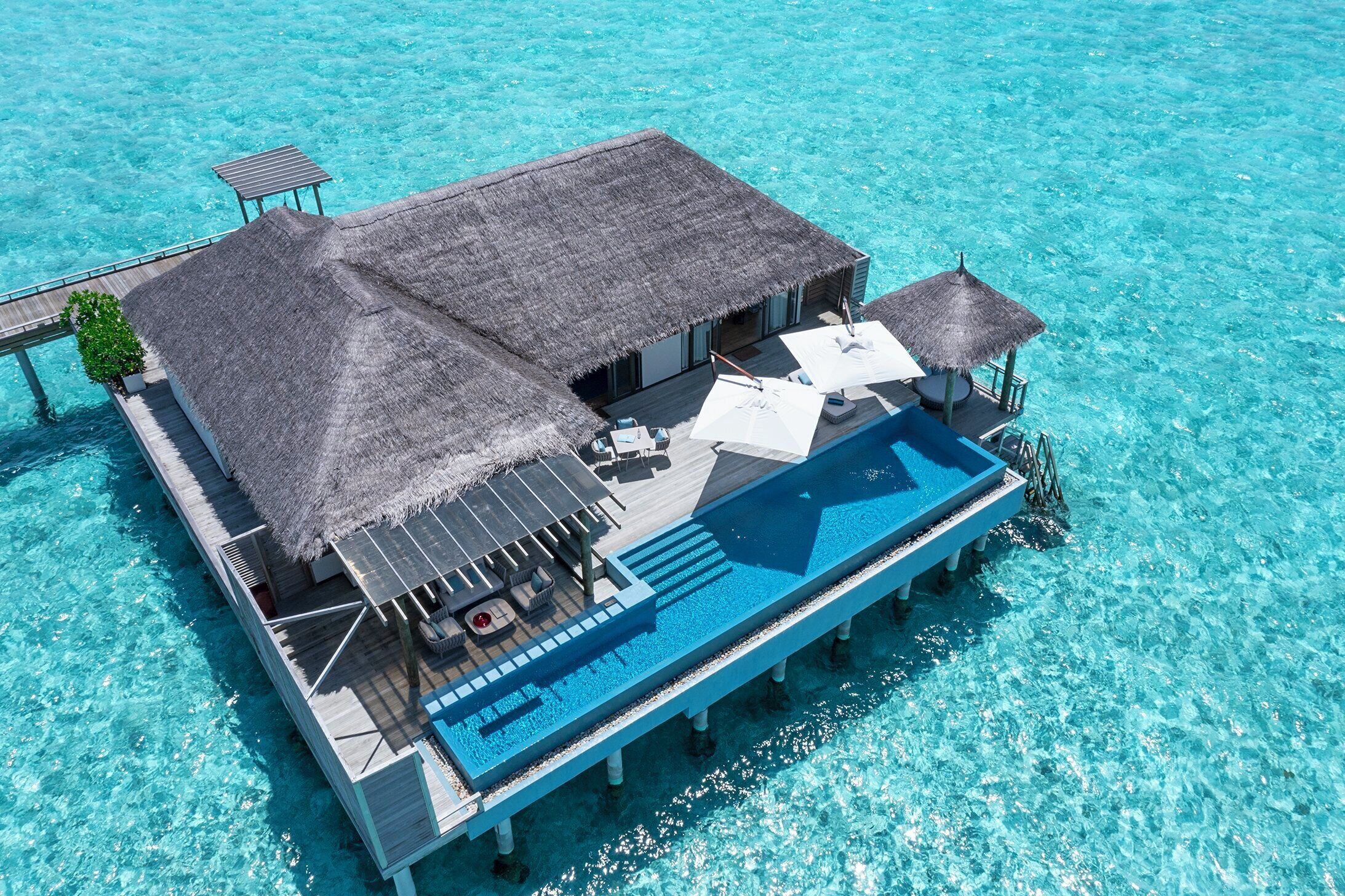 An overwater bungalow with a thatched roof, a swimming pool, and lounge chairs.