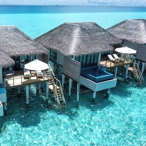 Cabañas sobre el agua con techos de paja, agua azul cristalina y muebles de terraza de madera.