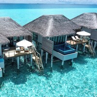 Cabañas sobre el agua con techos de paja, agua azul cristalina y muebles de terraza de madera.