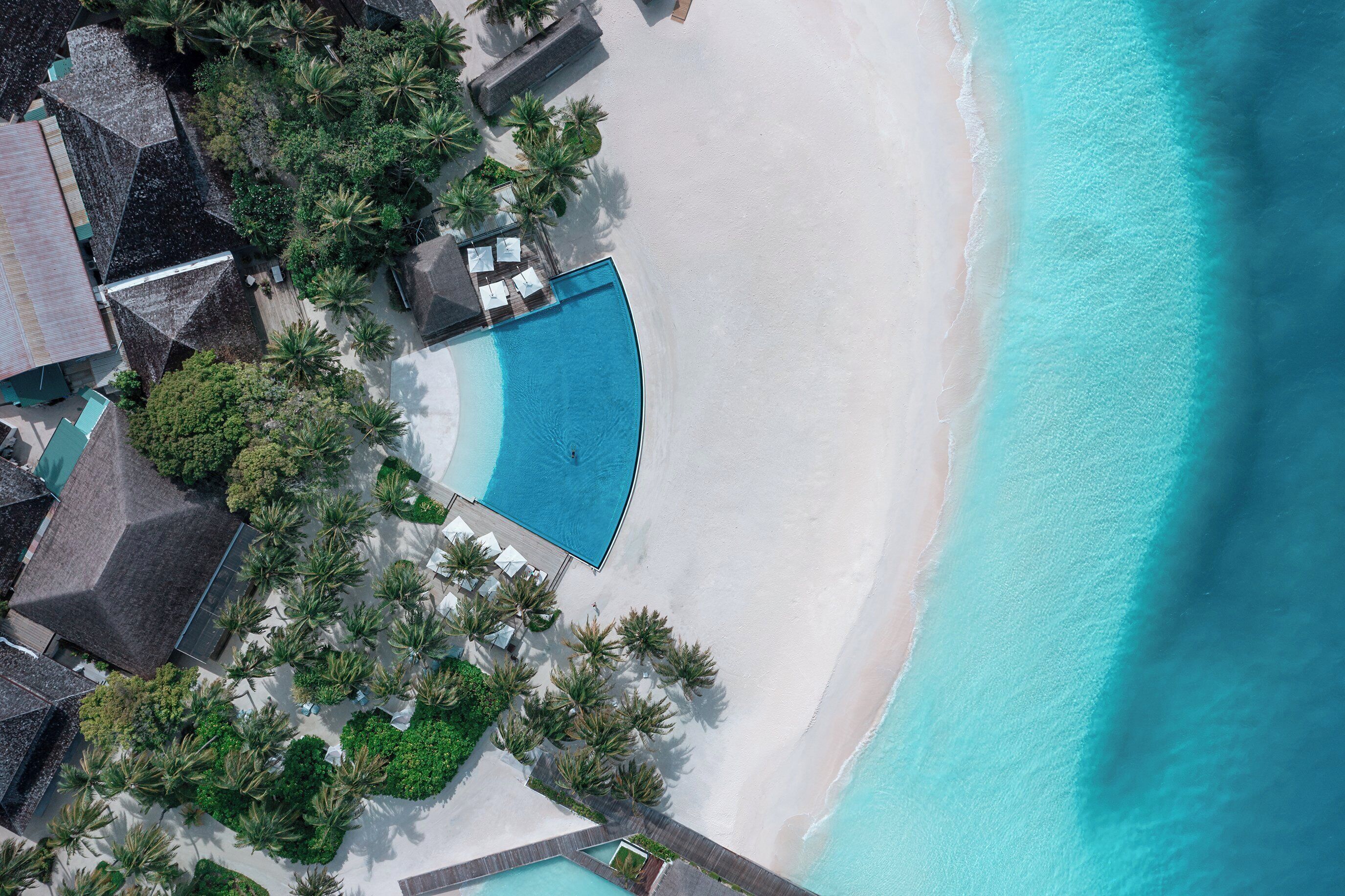 An aerial view of a resort with a swimming pool, beach, and palm trees.