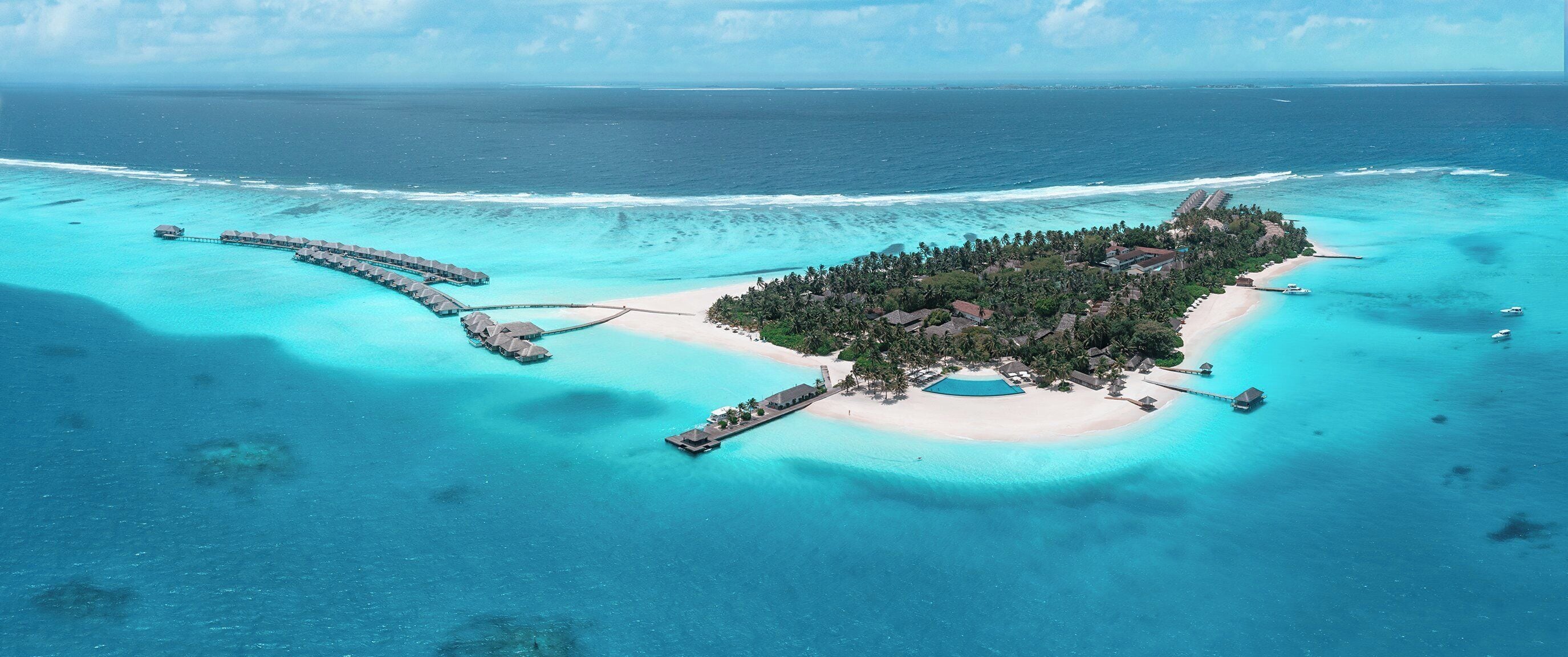Vista aérea de una isla tropical con bungalós sobre el agua conectados por pasarelas, una playa y aguas turquesas cristalinas.