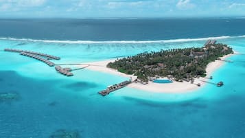 Vista aérea de una isla tropical con bungalós sobre el agua conectados por pasarelas, una playa y aguas turquesas cristalinas.