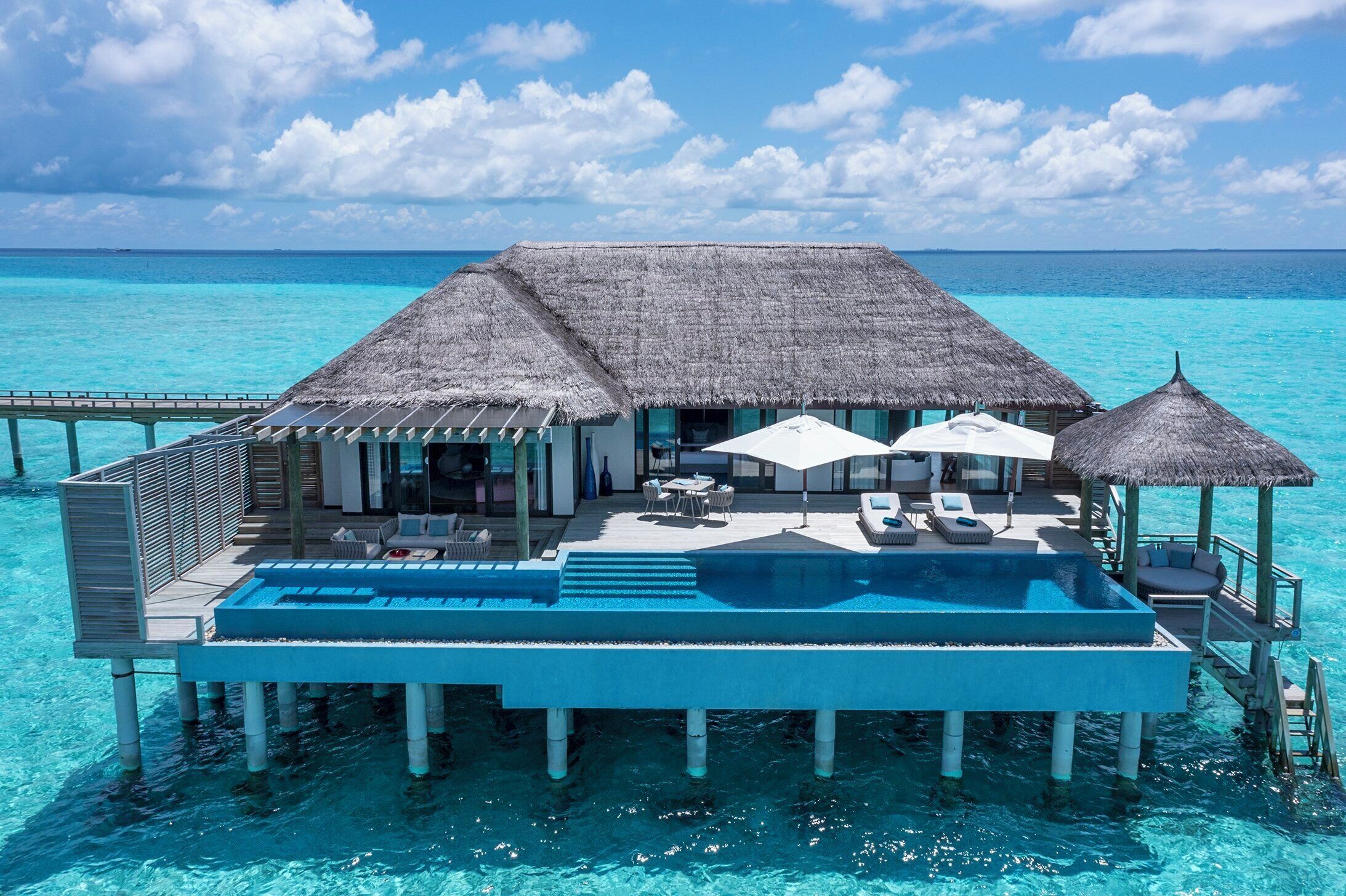 An overwater villa with a swimming pool, thatched-roof structures, and a view of the ocean.