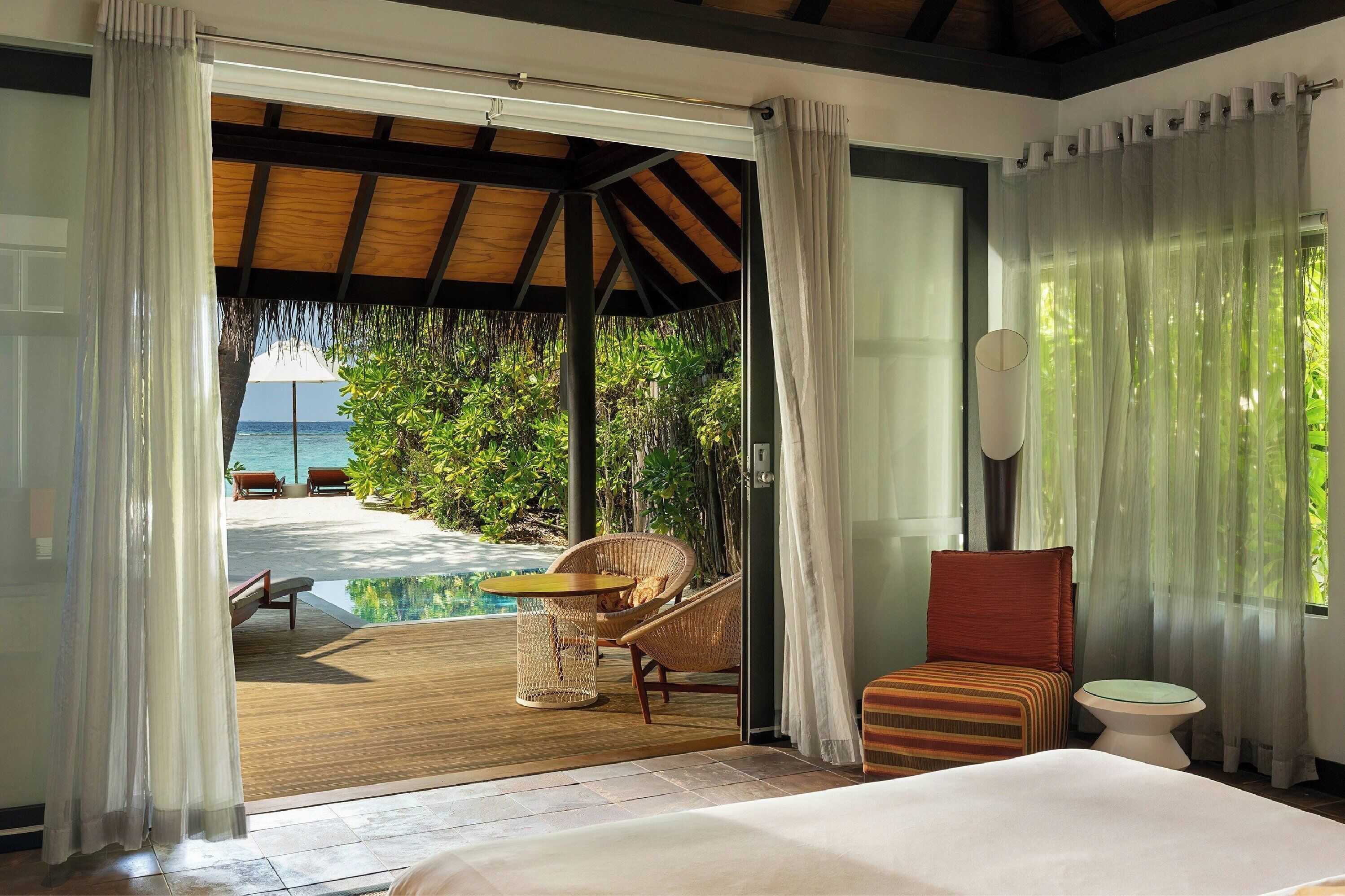 A room with a bed, a small table, a chair, and a toilet. There is a view of a pool and a beach.
