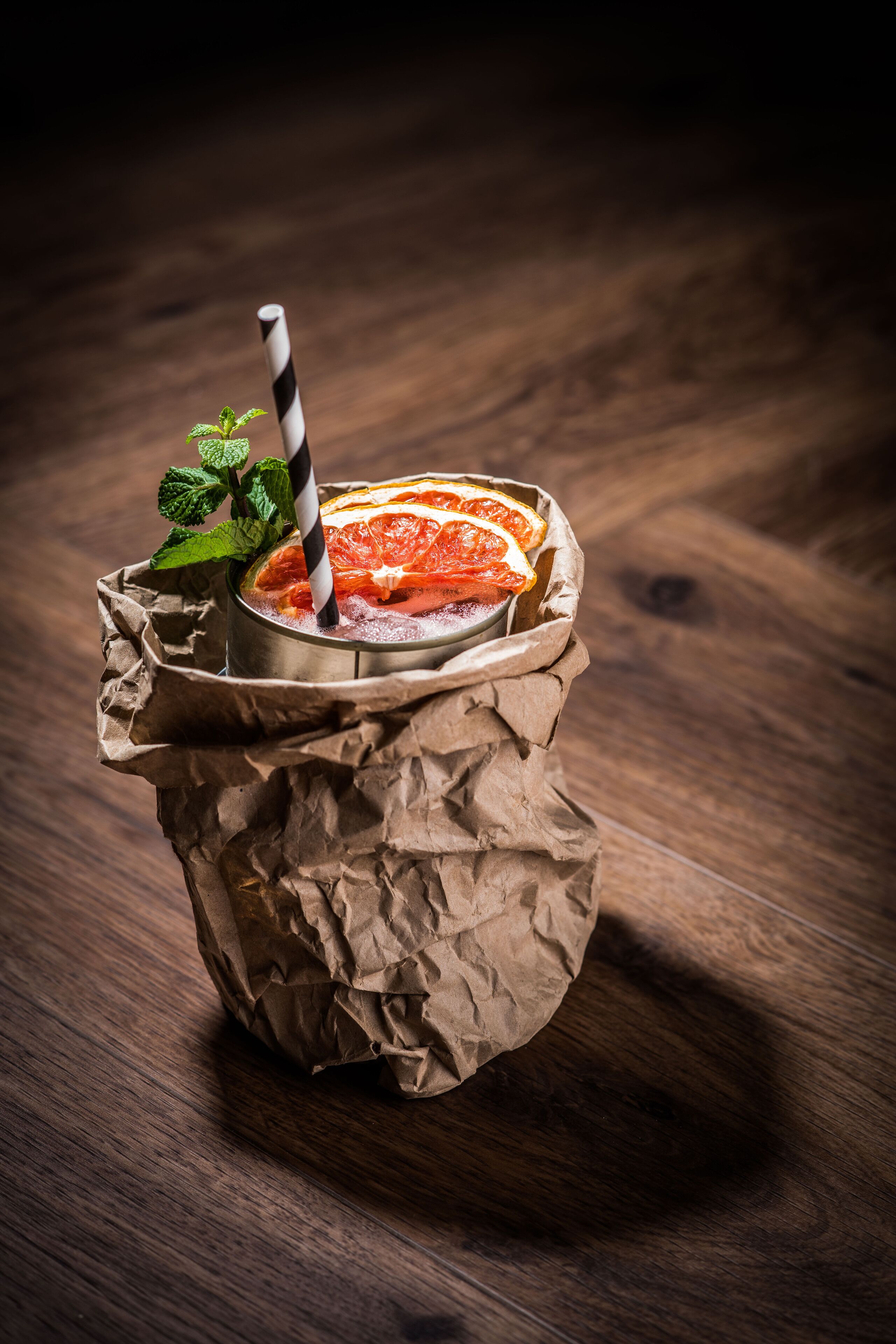A drink in a crumpled paper cup with a striped straw, garnished with mint and orange slices.