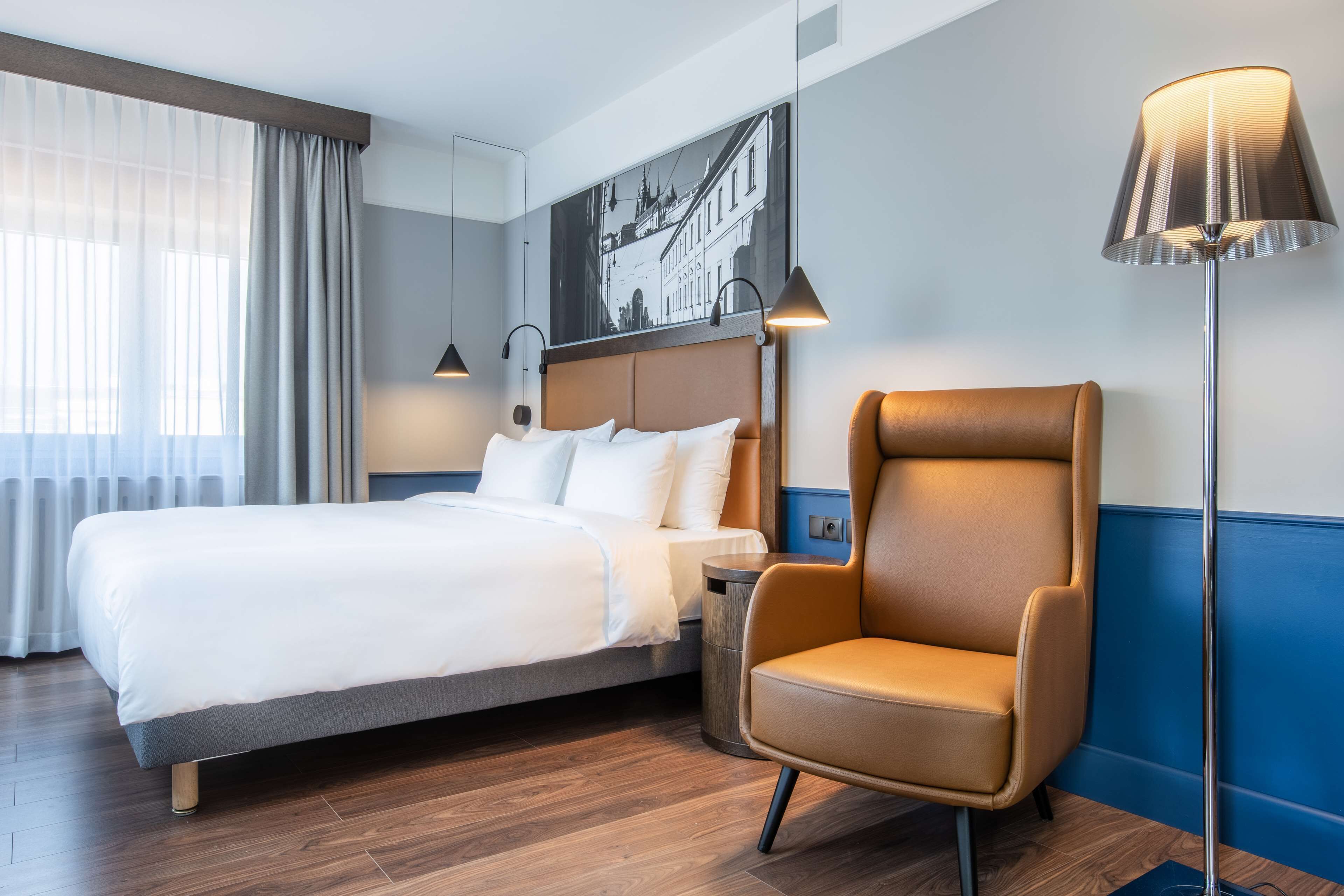 Una habitación de hotel con una cama, una silla, una lámpara de pie y un cuadro colgado en la pared.