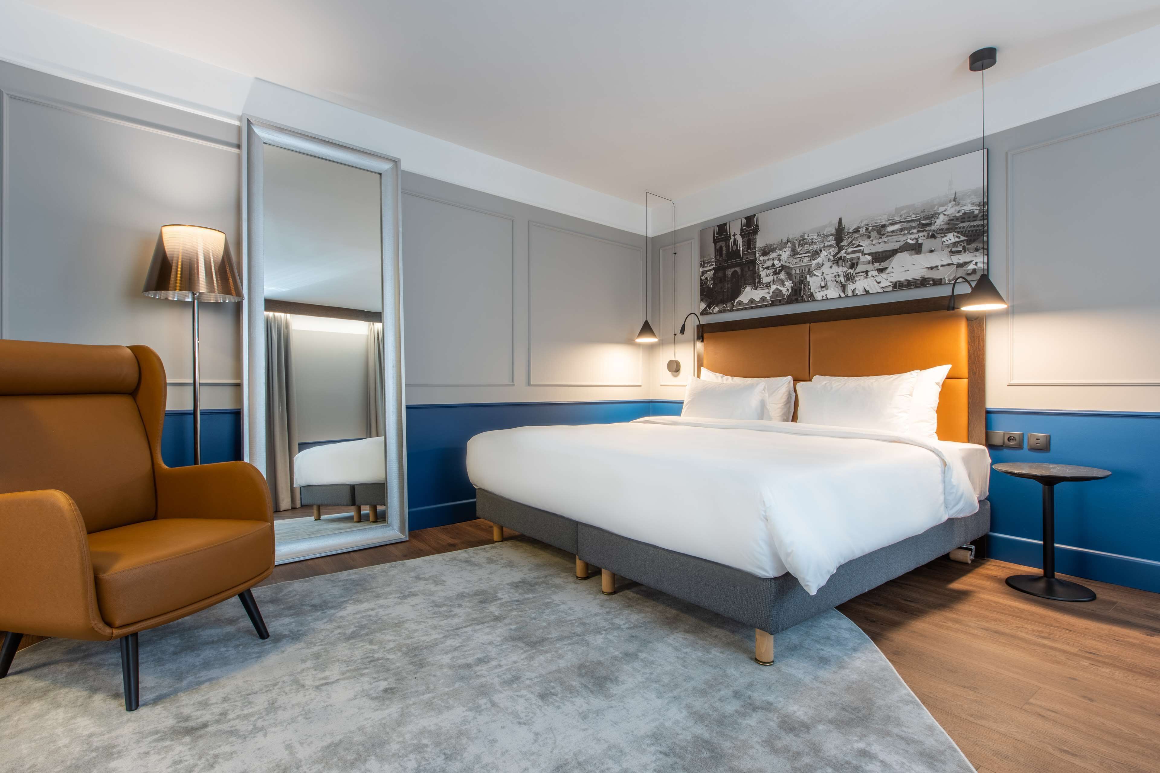 Una habitación de hotel moderna con una cama grande, una silla, una mesita y un espejo amplio.