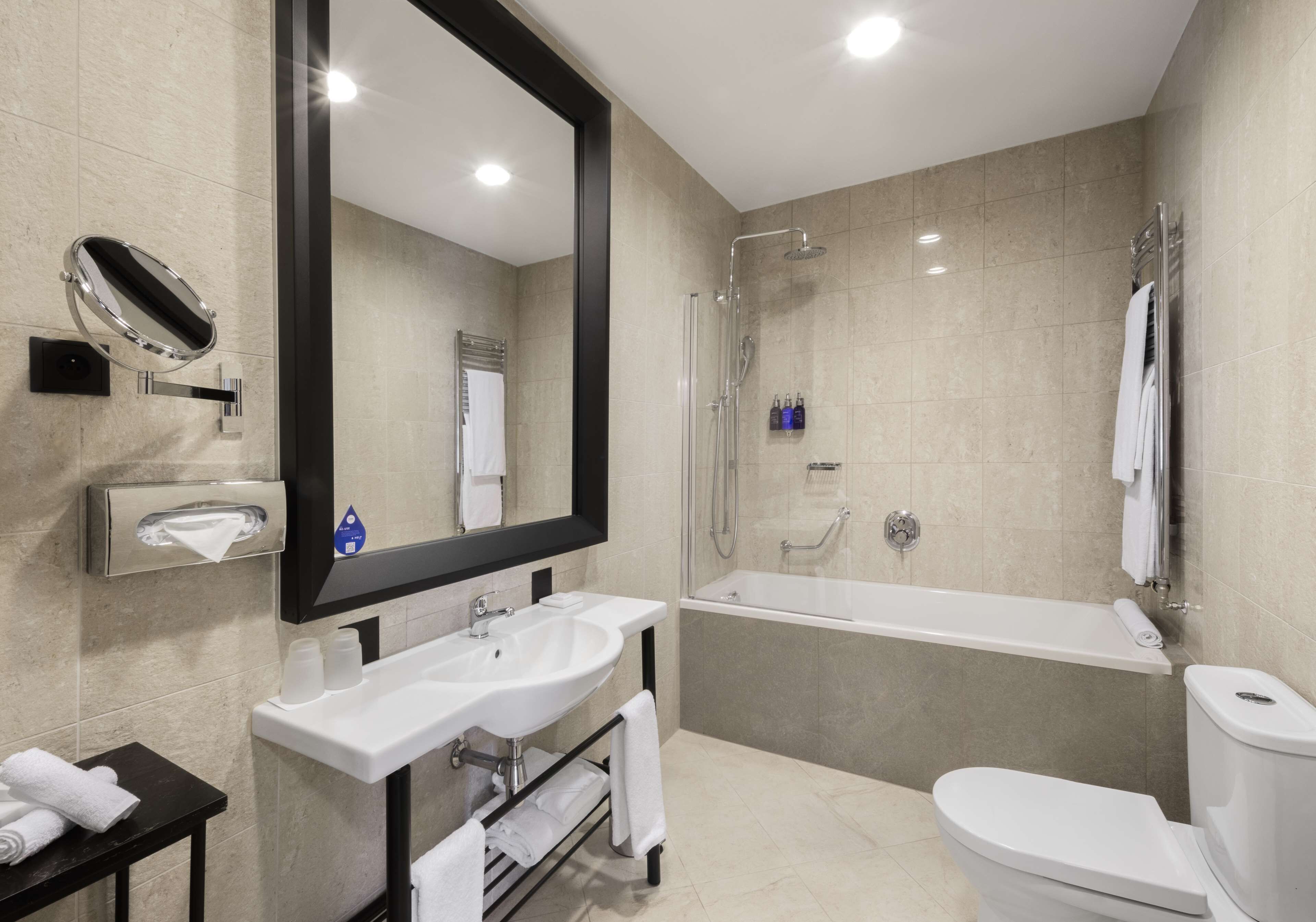 A modern bathroom with a large mirror, two sinks, a bathtub, and a toilet.