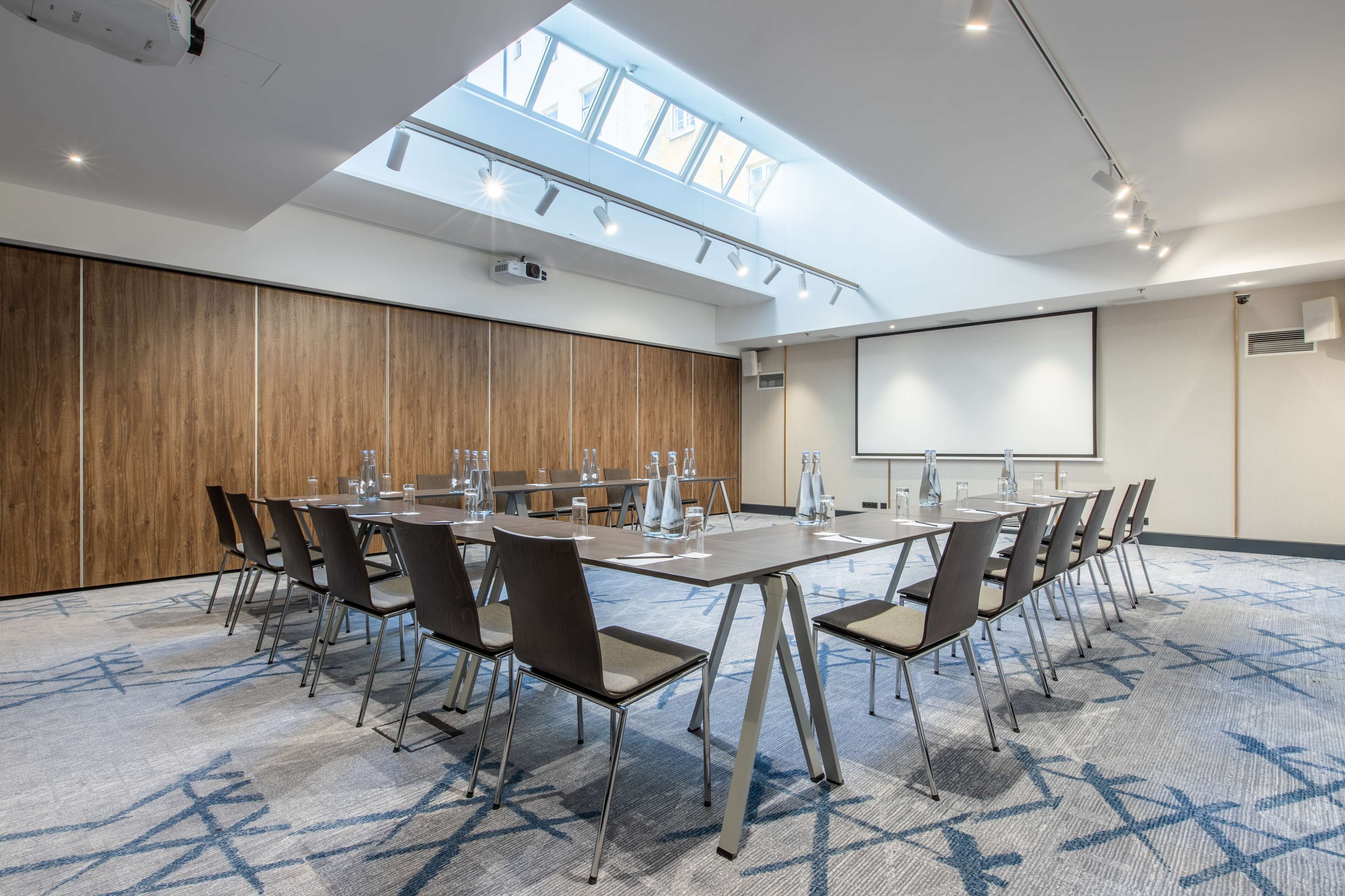 Un salón de conferencias con una mesa larga, sillas, una pantalla de proyector y un tragaluz.