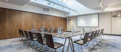 Un salón de conferencias con una mesa larga, sillas, una pantalla de proyector y un tragaluz.