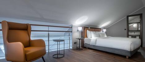 Una habitación de hotel moderna con un ventanal grande, un sillón de cuero marrón, una cama con ropa blanca, una mesita redonda y una lámpara de noche.