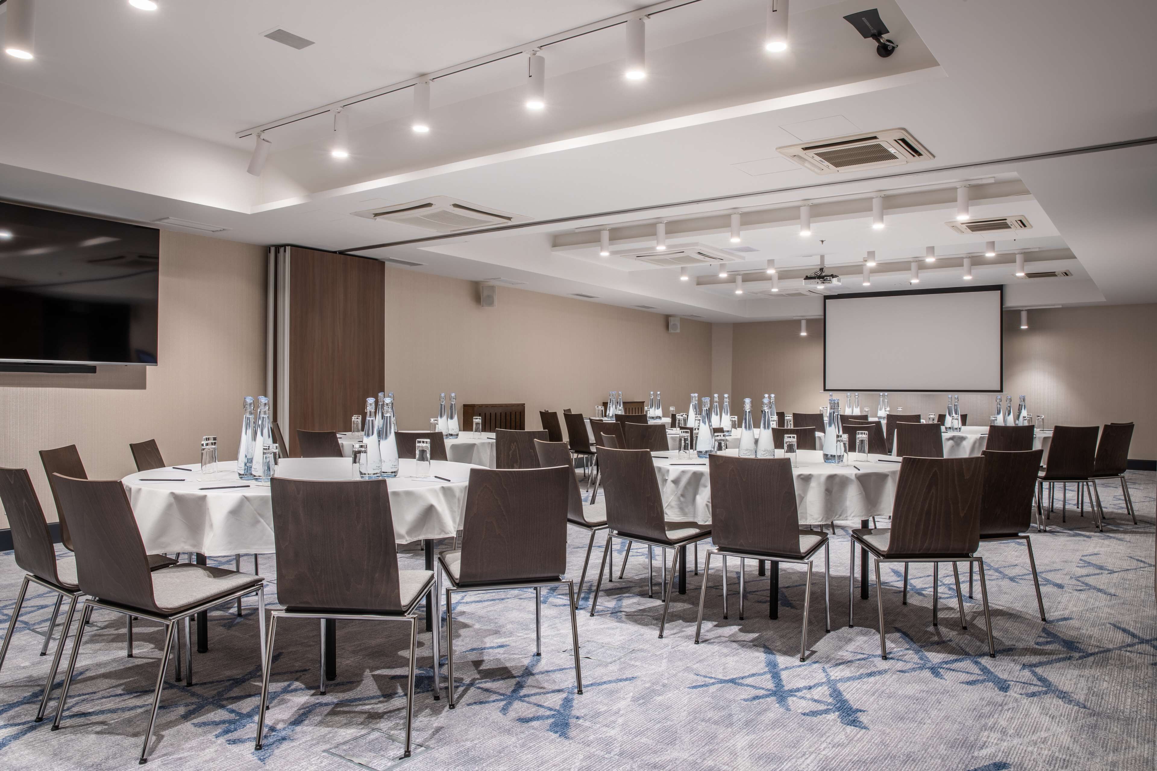 A conference room with multiple tables, chairs, and a large screen.