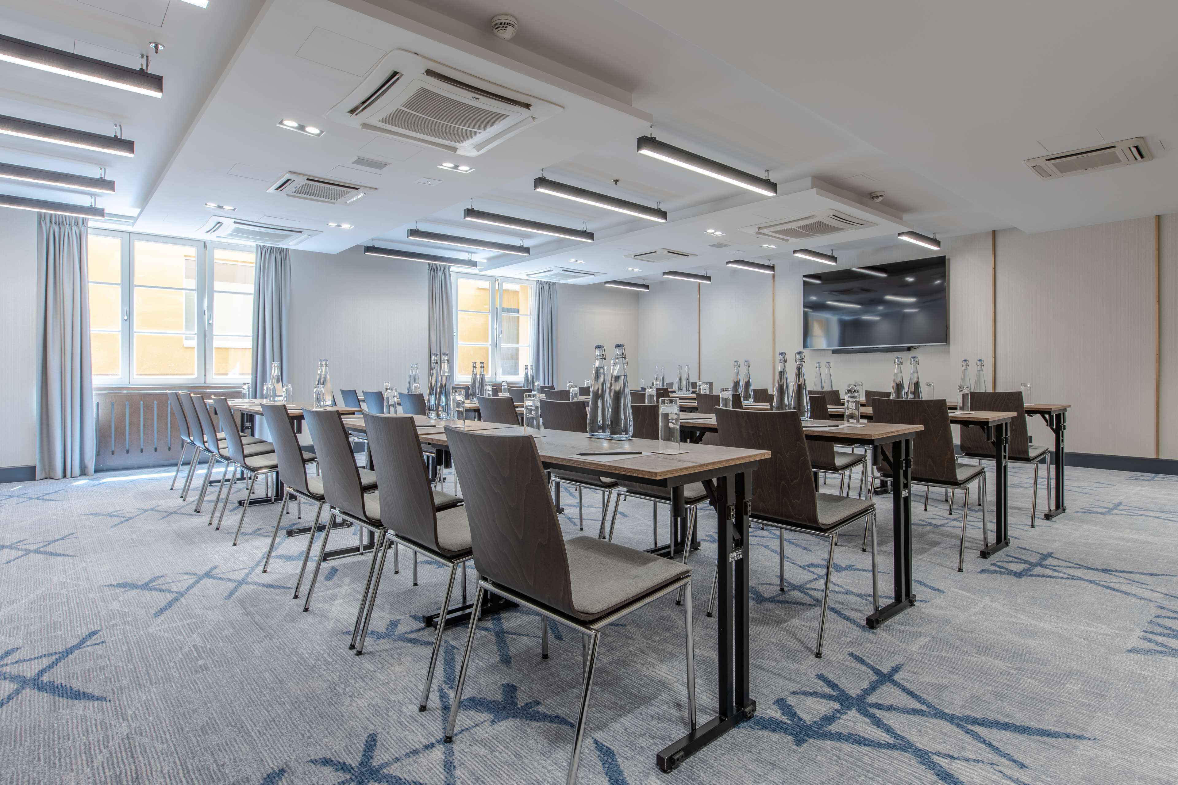 A conference room with multiple tables and chairs, large windows with curtains, and a flat-screen TV on the wall.