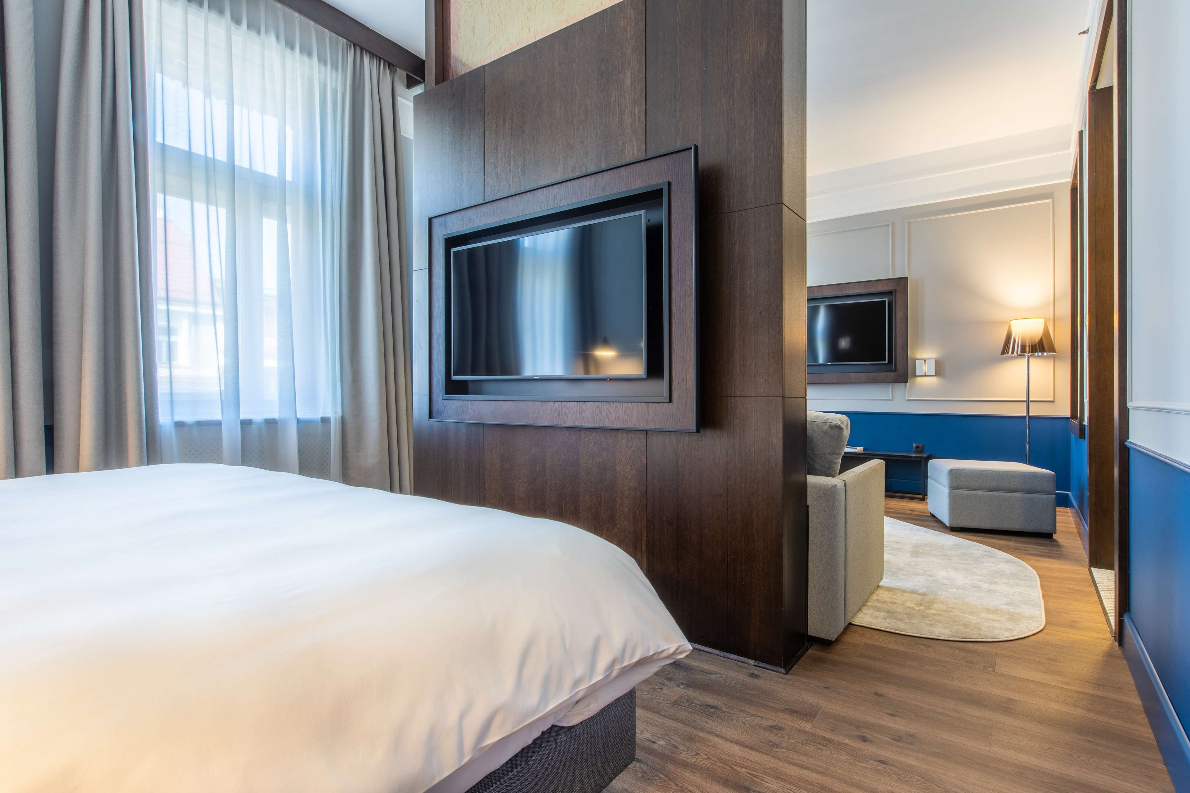 A modern hotel room with a large bed, a flat-screen TV mounted on the wall, a seating area with a sofa and ottoman, and a small table.