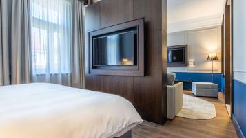 A modern hotel room with a large bed, a flat-screen TV mounted on the wall, a seating area with a sofa and ottoman, and a small table.