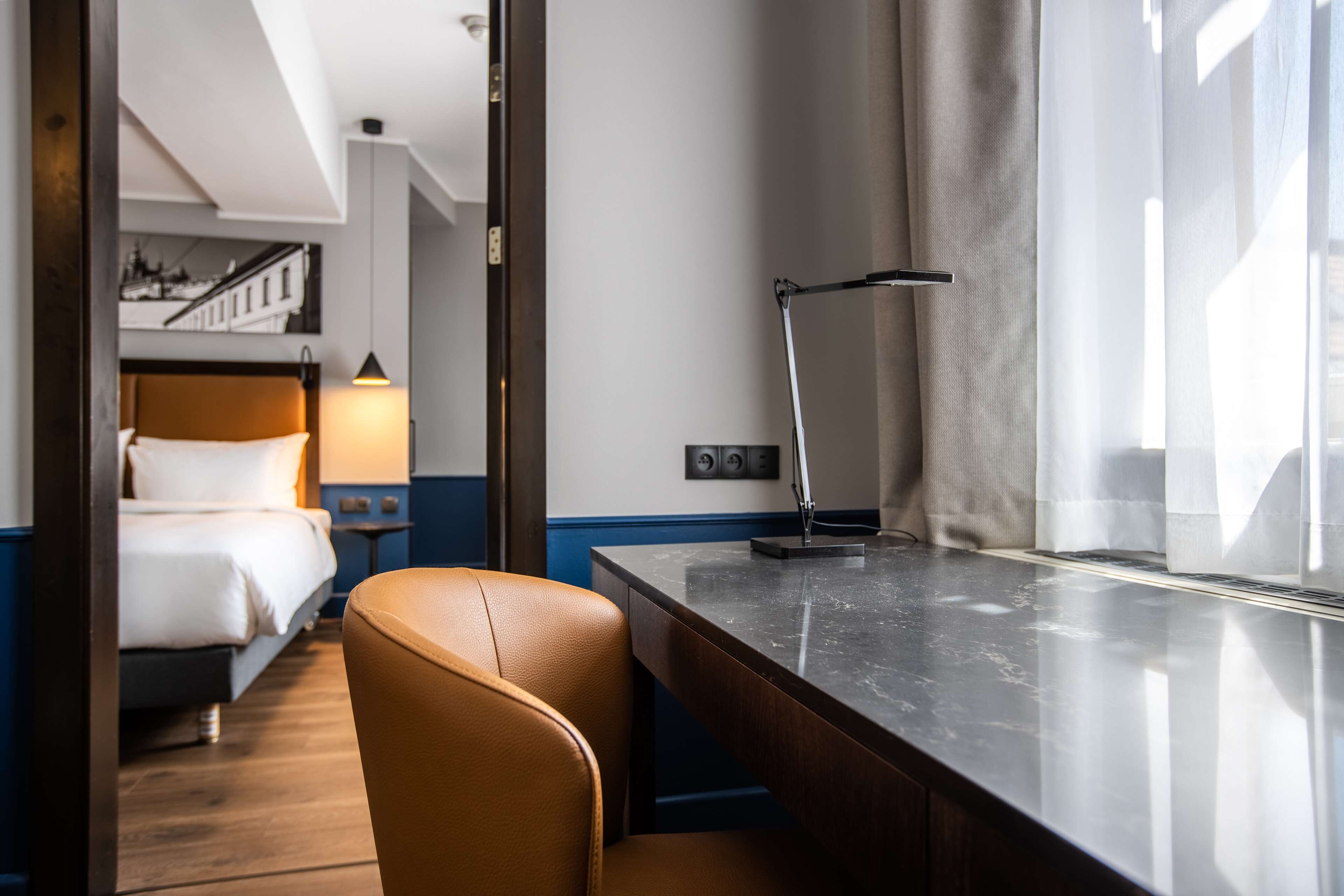Habitación de hotel con cama, escritorio, silla, lámpara, ventana con cortinas y vista a un edificio.