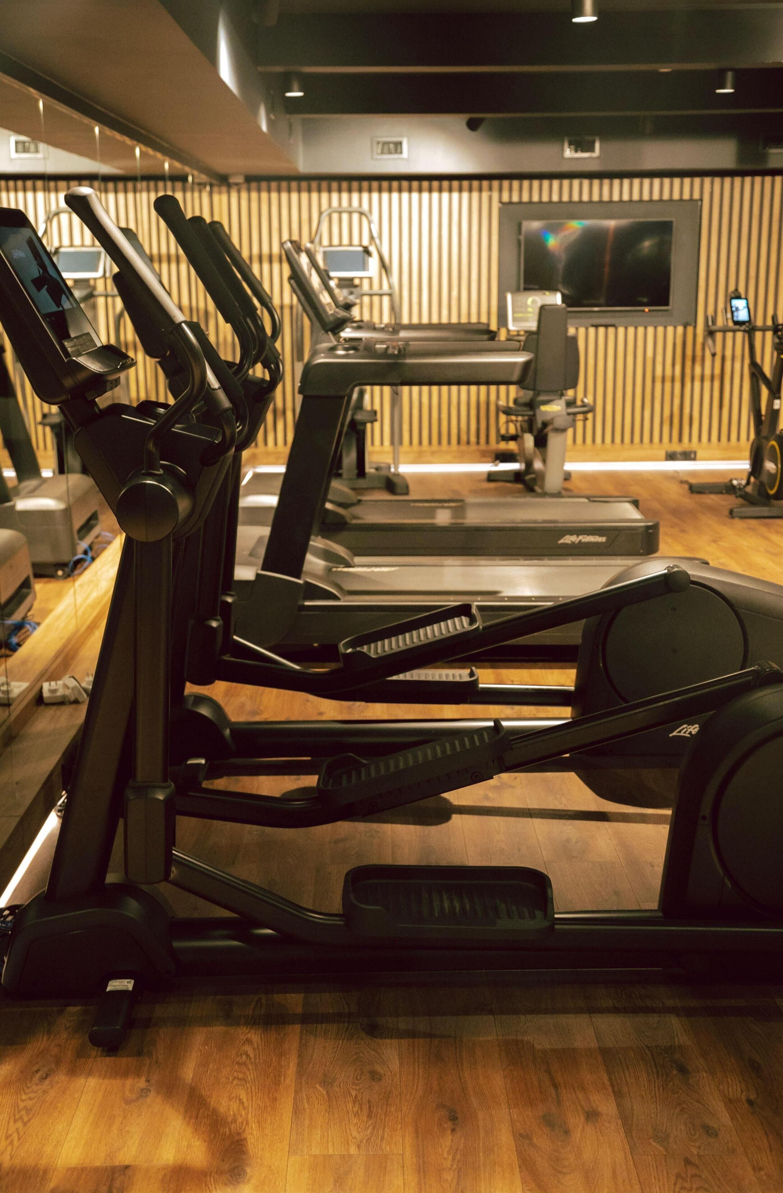 A gym with treadmills, stationary bikes, and a television screen on the wall.