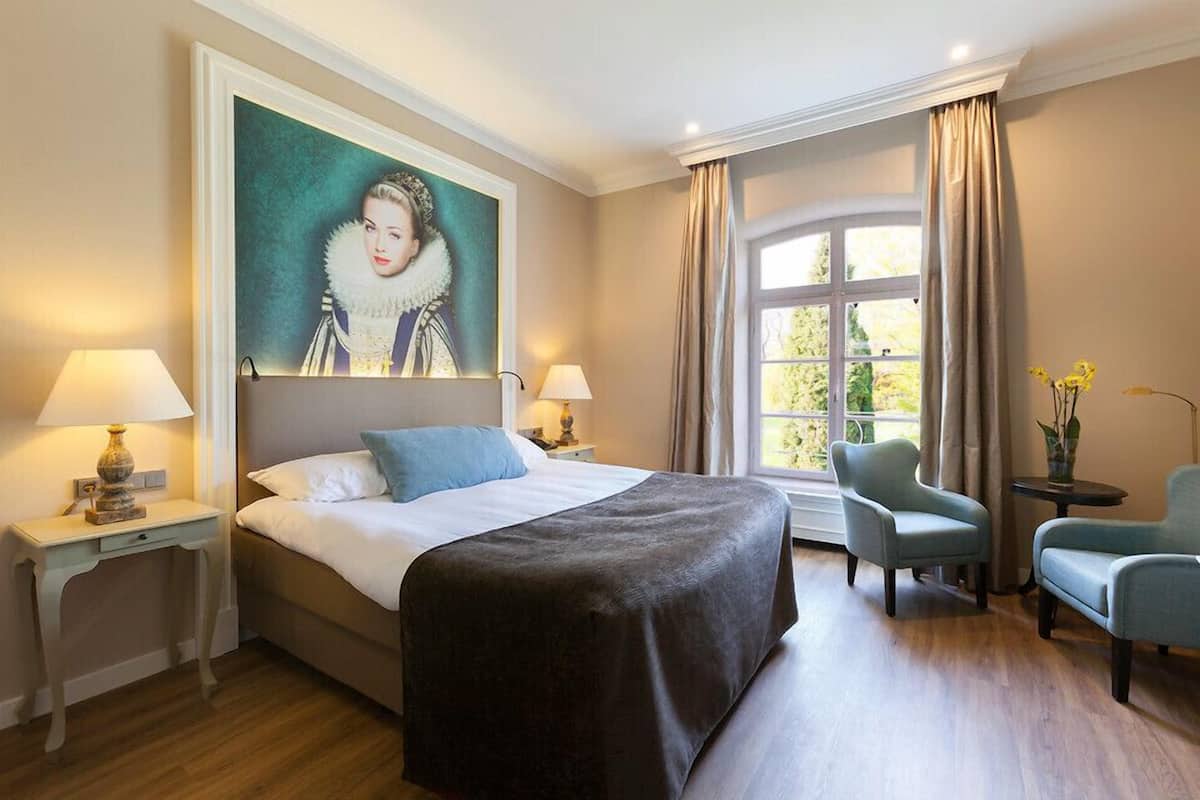 Habitación de hotel con una cama grande, dos mesitas de noche con lámparas, una silla, una pequeña mesa con un florero y un ventanal con cortinas.