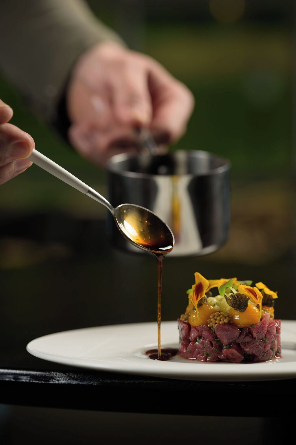 A spoonful of sauce being poured onto a dish of steak tartare with garnishes.
