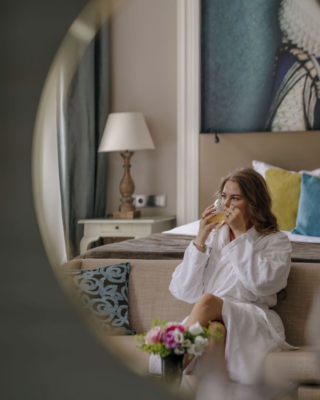 A person sitting on a sofa in a room with a lamp, a bed, and a painting on the wall.