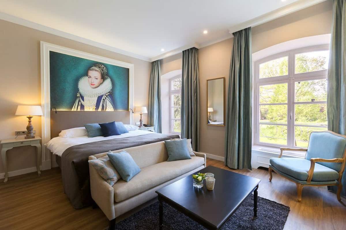 Habitación de hotel con una cama grande, un sofá, una mesa de centro, una silla, un cuadro y un ventanal con cortinas.