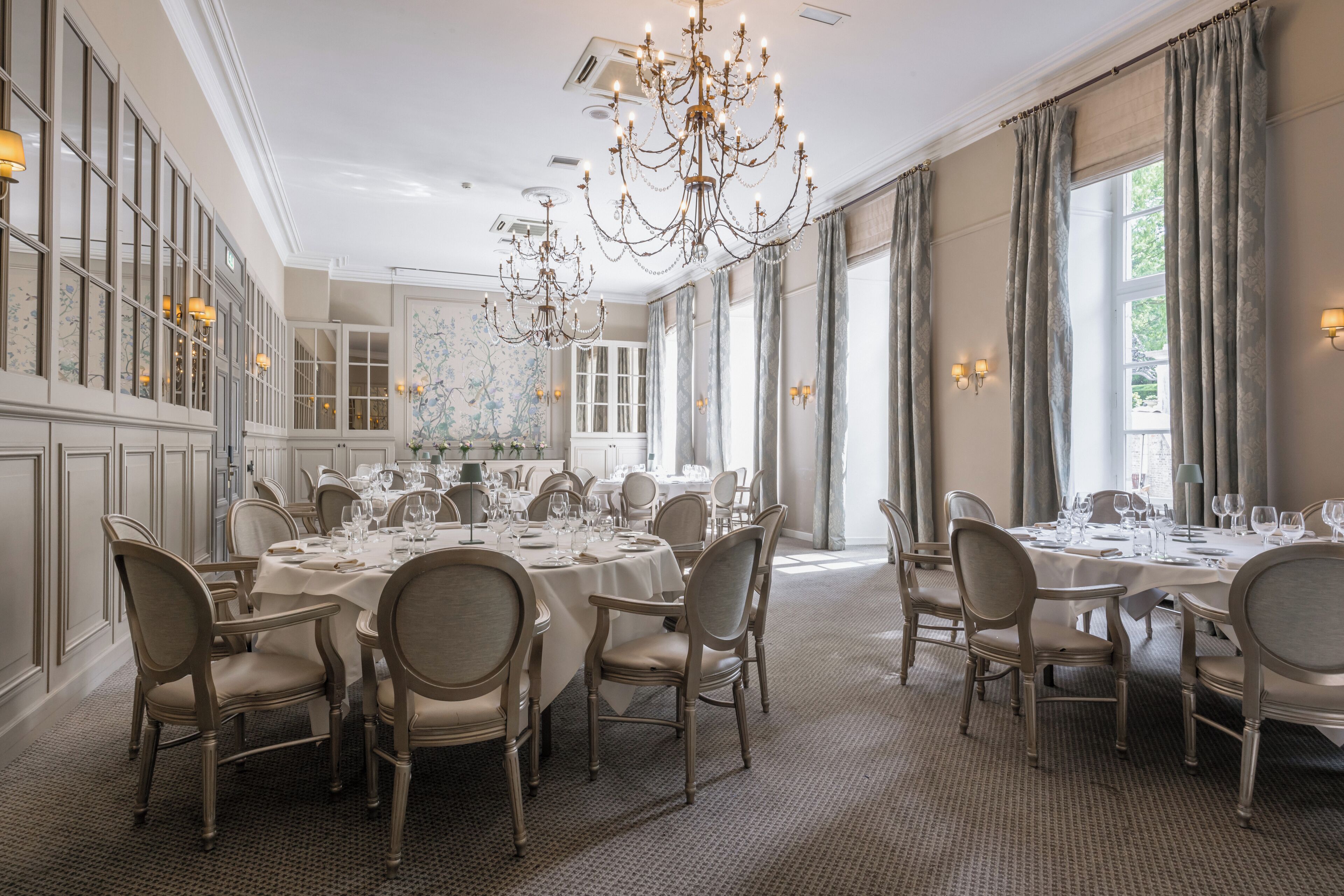 Uma sala de jantar formal com várias mesas redondas preparadas para o serviço, lustres elegantes e grandes janelas com cortinas.