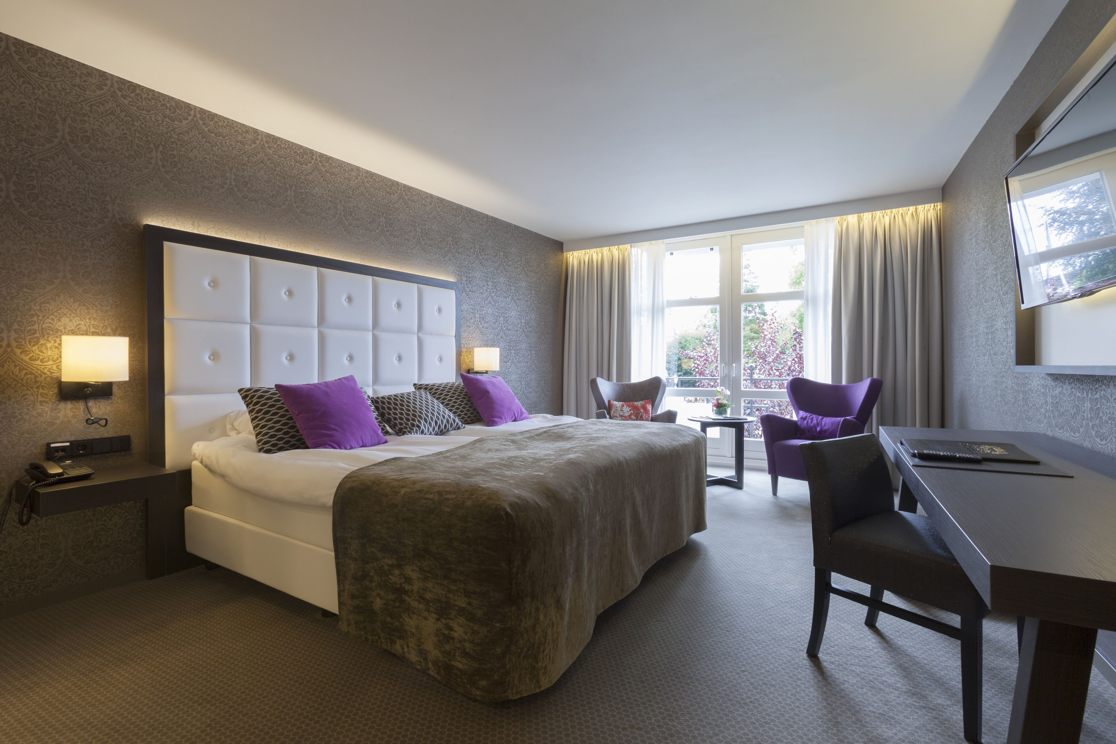 Een hotelkamer met een groot bed, twee stoelen, een bureau en een raam met gordijnen.