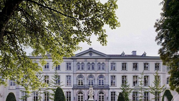 Un edificio grande de estilo clásico con arquitectura simétrica, una estatua central y un jardín bien cuidado con fuentes.