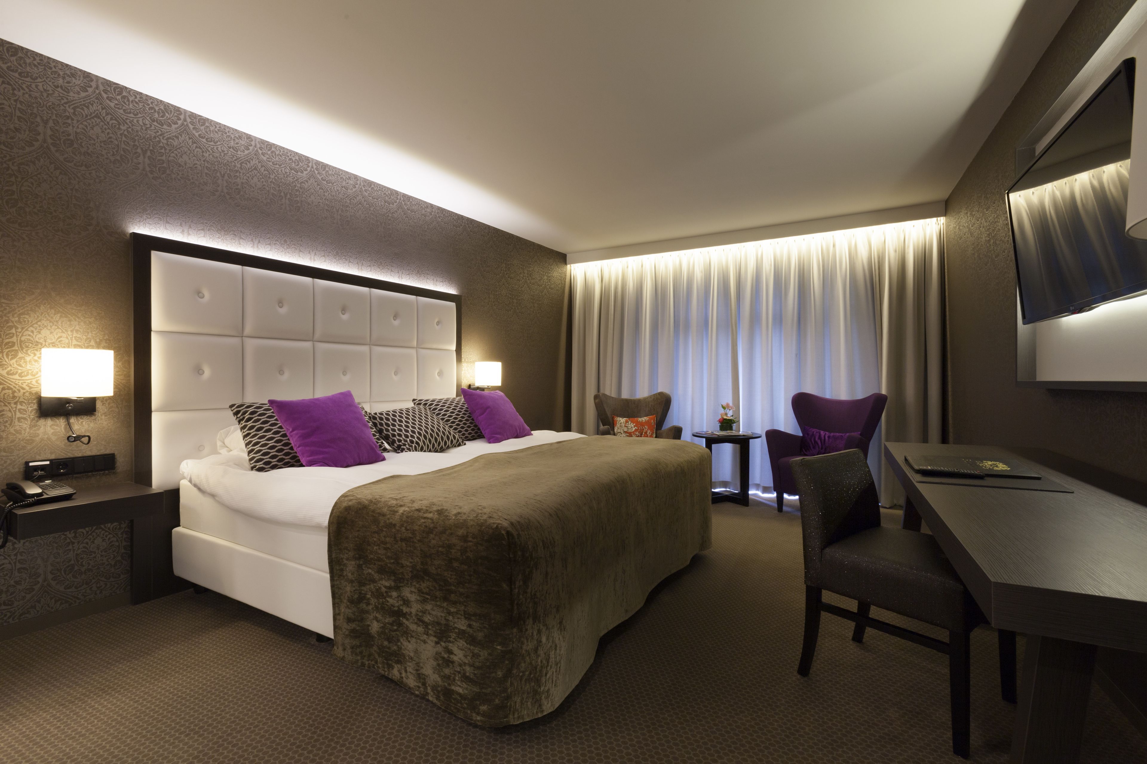 A hotel room with a large bed, a desk, a chair, a nightstand, and a lamp.